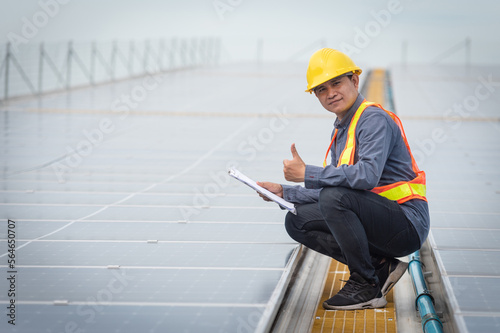 Portrait of Asian electrician engineer in safety helmet and uniform using laptop checking solar panels. Male technician at solar station.