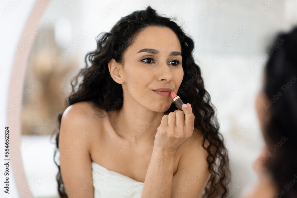 Glad pretty islamic millennial curly female in towel applying lipstick on lips, looking in mirror in bedroom
