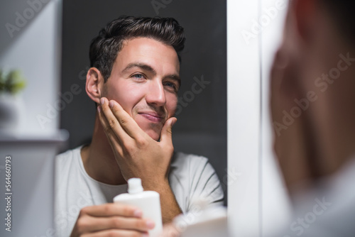 Man is applying aftershave while looking himself in the mirror.	
