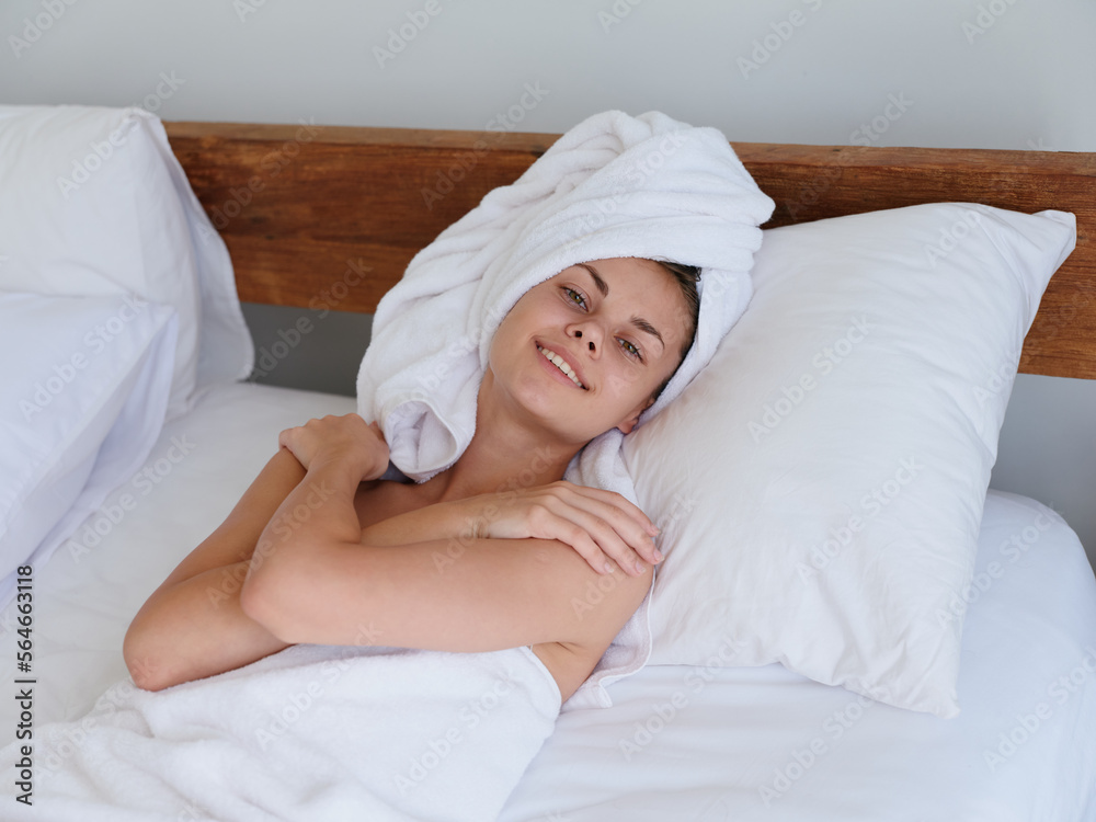 A woman lies on the bed after a shower with wet hair with a towel on her head in the bedroom and strokes her body skin, a cute smile with teeth and weekend rest in bed, women's beauty day, lifestyle A woman lies on the bed after a shower with wet hair with a towel on her head in the bedroom and strokes her body skin, a cute smile with teeth and weekend rest in bed, women's beauty day, lifestyle