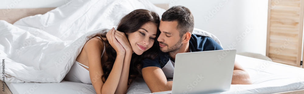 Young man in pajama looking at brunette girlfriend near laptop on bed, banner. Young man in pajama looking at brunette girlfriend near laptop on bed, banner.