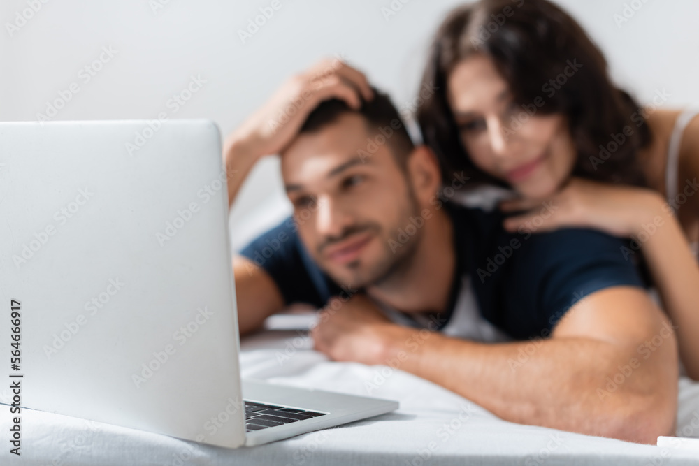 Laptop near blurred couple lying on bed at home.