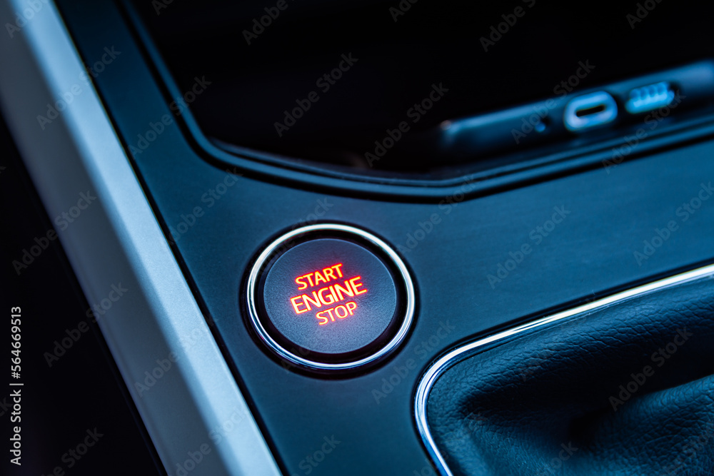 Close-up view of round shaped start engine button on the car interior, highlighted in red, on center console made from dark plastic with silver frames. Blurred mobile phone on the background. 