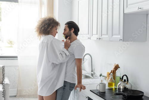 young woman in shirt seducing bearded man in jeans in kitchen.