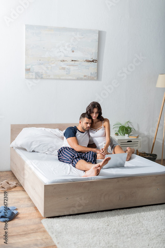 Couple in pajama holding hands near laptop on bed in morning.