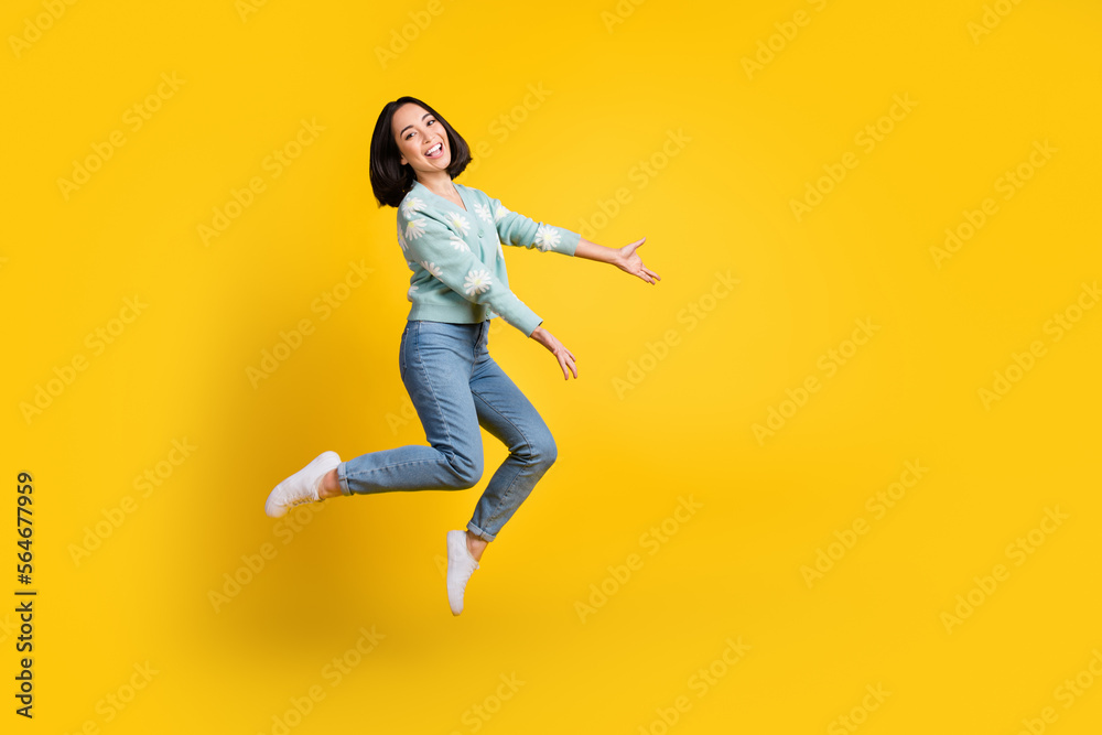 Full length photo of cheerful girl dressed blue cardigan jeans jump up arms demonstrate empty space isolated on yellow color background