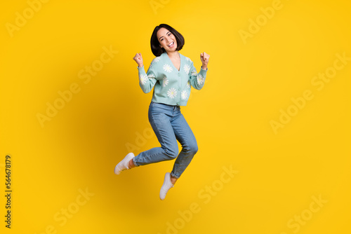 Full length photo of girlish nice satisfied girl wear blue cardigan jeans clenching fists jumping isolated on yellow color background