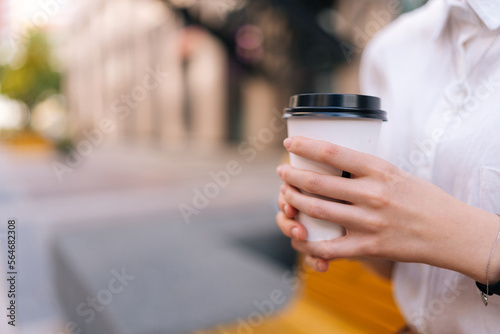 Close-up cropped shot of unrecognizable young woman holding in hands paper cup with takeaway coffee sitting on bench on city street, in summer sunny day on blurred background, selective focus.