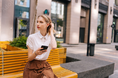 Portrait of charming young woman with long blond hair holding mobile phone in hand while sitting on yellow bench on city street and looking away. Lady spending time in urban summer street using gadget