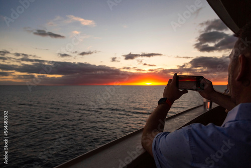 Man taking a smartphone photo of a beautiful sunset while enjoying an evening on a luxury cruise ship. Cruise vacations are relaxing and enjoyable photo