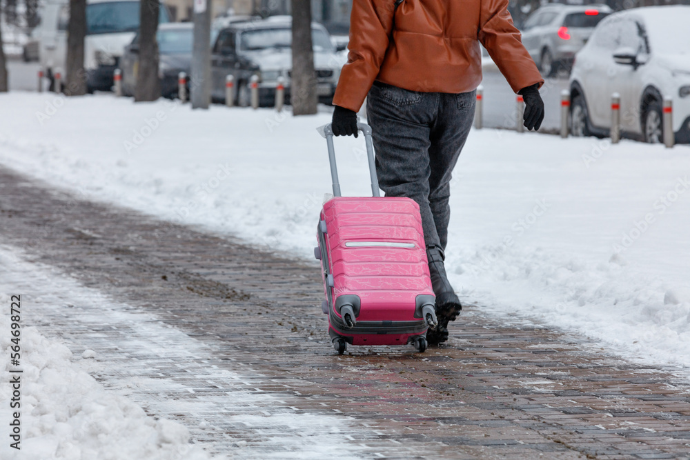 A traveler woman walks along a paved sidewalk in the winter season, carrying a travel bag on wheels. Copy space. A traveler woman walks along a paved sidewalk in the winter season, carrying a travel bag on wheels. Copy space.