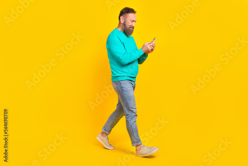 Full length photo of nice retired man walking hold device reading news wear trendy aquamarine clothes isolated on yellow color background