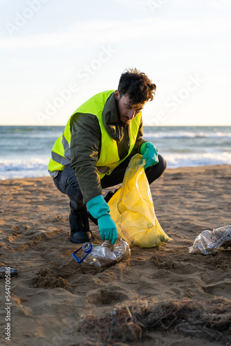 Volunteer bending down picking up plastic jug from the sand on the beach. Vertical photo. Concept of caring for our planet