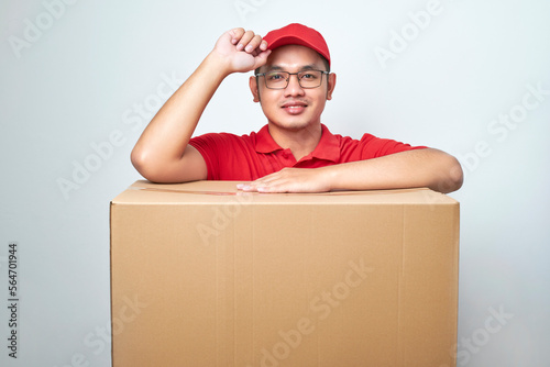 Friendly- ooking asian delivery guy in red cap and uniform, courier service lean on cardboard box or client package, saluring as making order photo