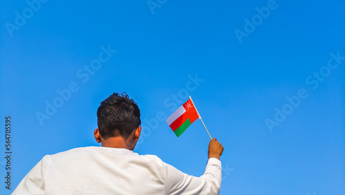 Boy holding Oman flag against clear blue sky. Man hand waving flag of Oman view from back, copy space