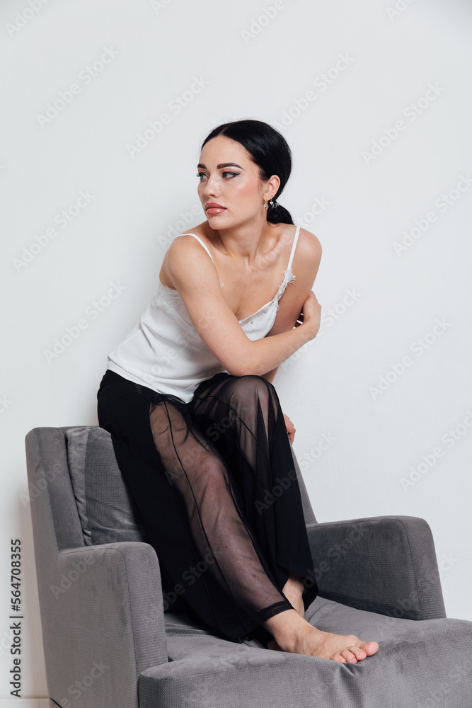 a fashionable woman in a white T-shirt and black pants sits on a chair with a white background