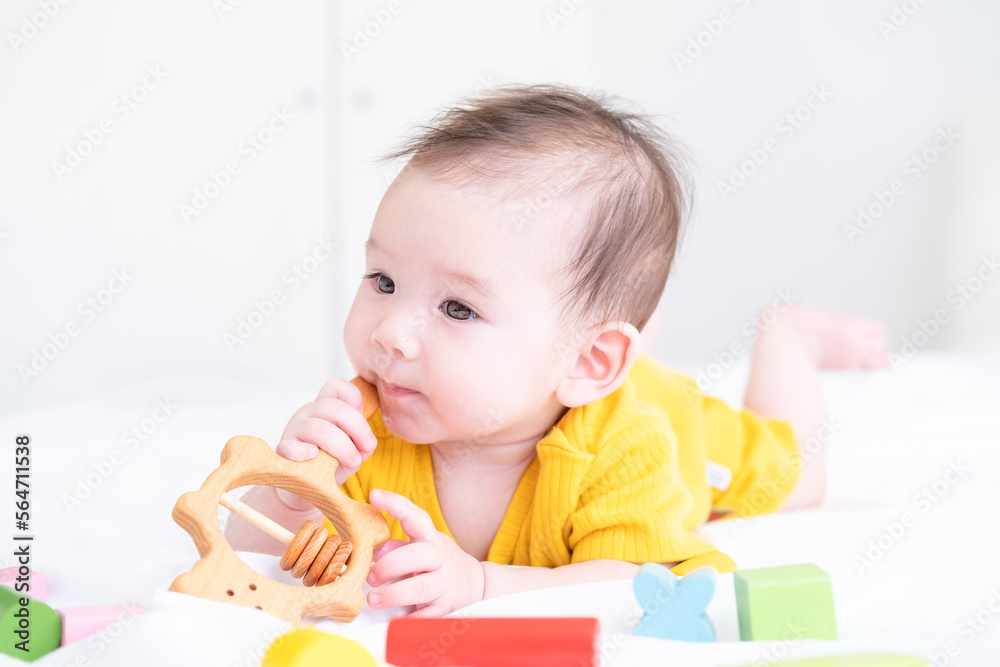 healthy asian baby girl in yellow bodysuit plays with wooden toy teether for teeth on white bedding healthy asian baby girl in yellow bodysuit plays with wooden toy teether for teeth on white bedding