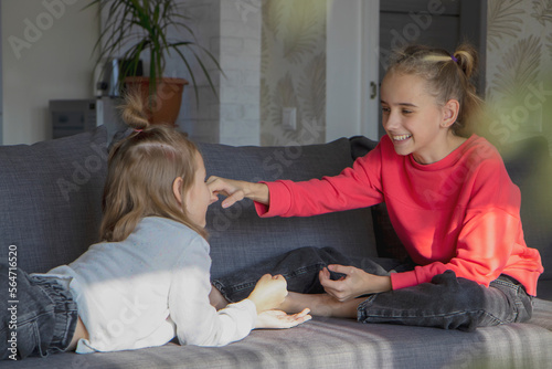 Two little sister girls play at home in the living room, look at each other, laugh and smile. The concept of warm relationships, love, trust and understanding in the family.