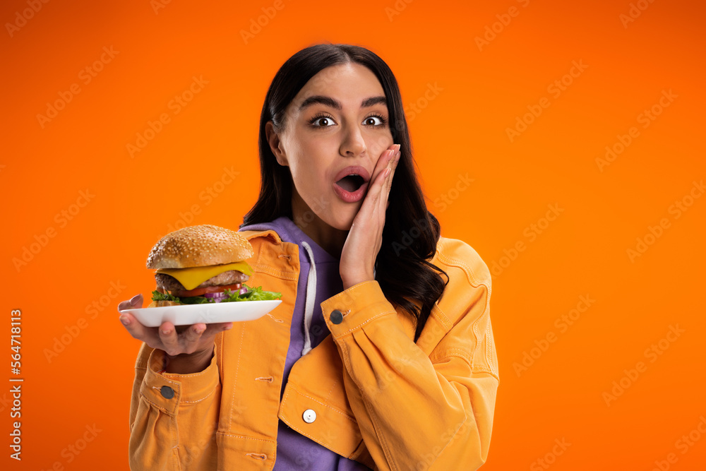 amazed woman in trendy jacket holding tasty burger while touching face and looking at camera isolated on orange. amazed woman in trendy jacket holding tasty burger while touching face and looking at camera isolated on orange.