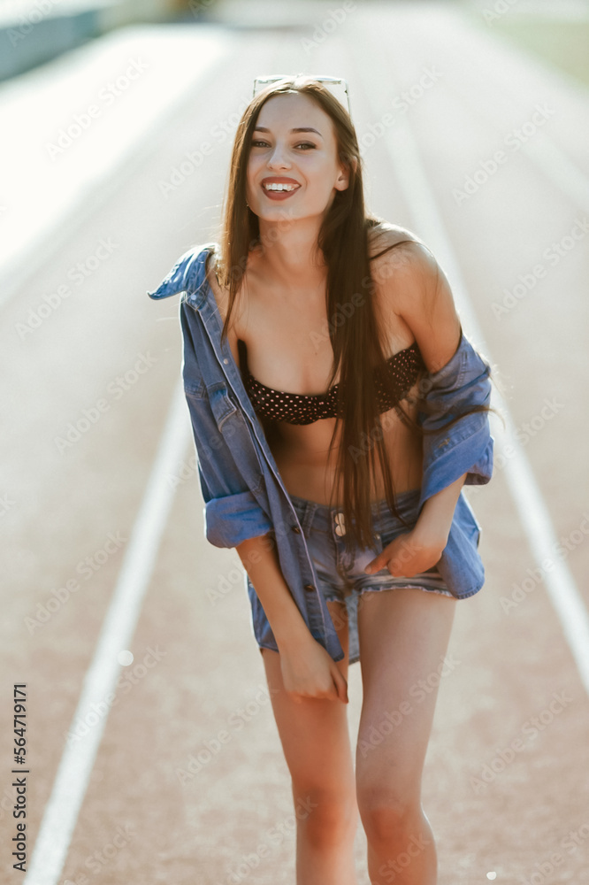 a teenage girl with long dark hair poses for the camera; on a stadium running track; a model-looking girl on the street; slender figure of a beautiful girl in a bikini and denim shorts;