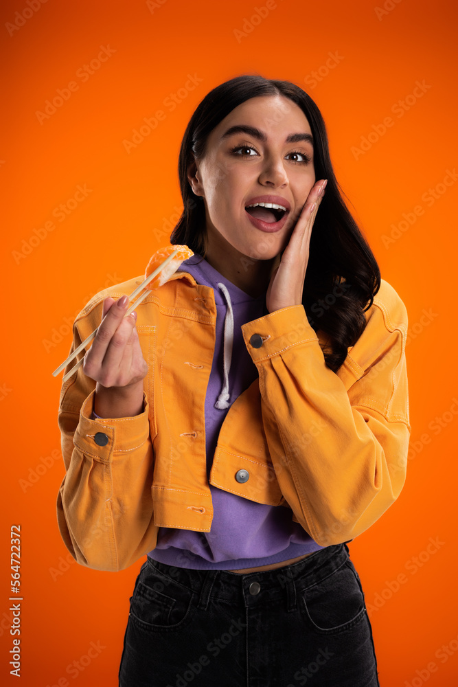 amazed woman holding delicious sashimi while touching face and looking at camera isolated on orange.