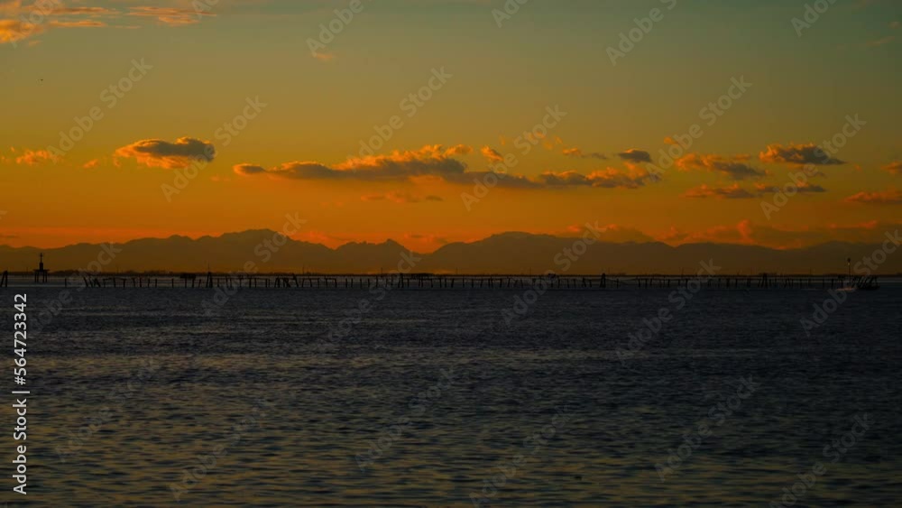 Blue rippling sea against distant high Alps mountains under breathtaking orange sky with massive cloud formations at sunset Blue rippling sea against distant high Alps mountains under breathtaking orange sky with massive cloud formations at sunset