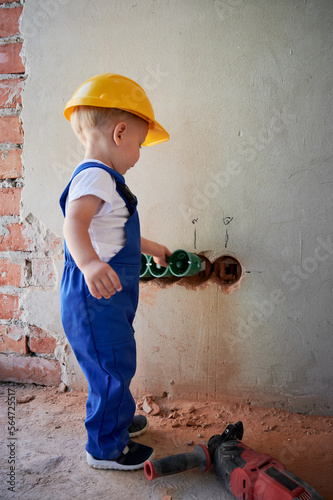 Little boy electrician installing cable canals and sockets in wall. Child in safety and work overalls construction helmet mounting electrical wiring in apartment under renovation.