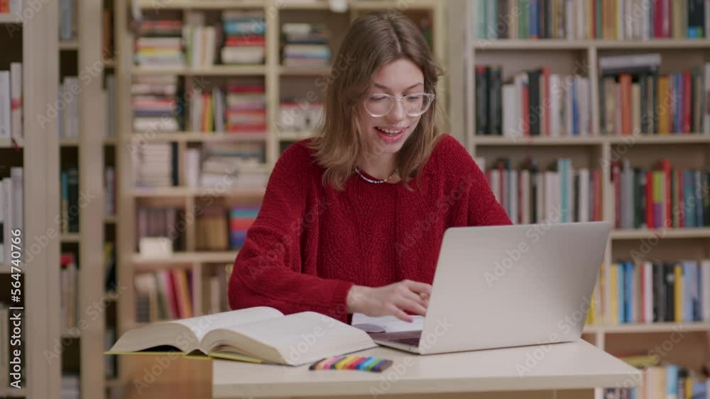 Smiling caucasian lady wearing red sweater and glasses work on laptop in library Smiling caucasian lady wearing red sweater and glasses work on laptop in library