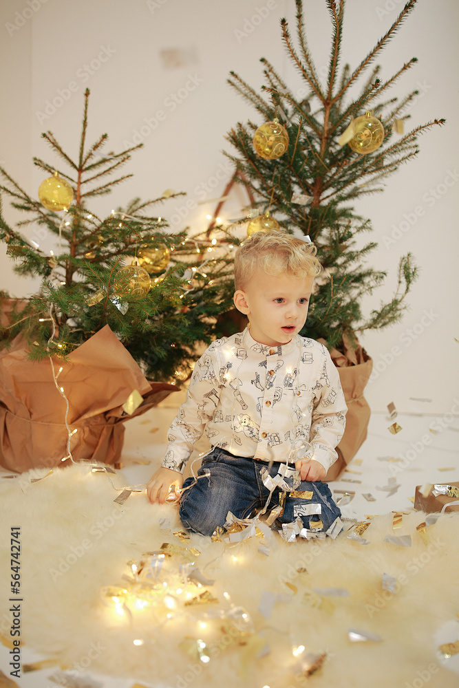 a little boy is sitting near a Christmas tree. Christmas children's photo session in the studio. New Year's photo zone in a photo studio. New Year's decor. a little blond boy with blue eyes a little boy is sitting near a Christmas tree. Christmas children's photo session in the studio. New Year's photo zone in a photo studio. New Year's decor. a little blond boy with blue eyes