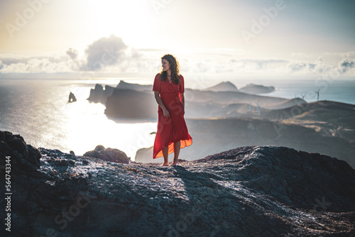 Woman in red dress enjoys panoramic view from steep cliff over seascape and along rugged foothills of Madeira coast at sunrise. Ponta do Bode, Madeira Island, Portugal, Europe.