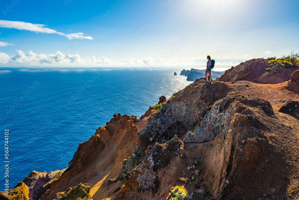 Backpacker woman enjoys view from a steep cliff overlooking the sea and the rugged foothills of Madeira's coast in the morning. Ponta do Bode, Madeira Island, Portugal, Europe.