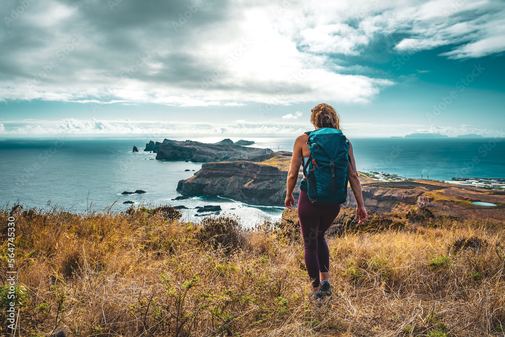 Backpacker woman enjoys hike along a steep cliff overlooking the sea and the rugged foothills of Madeira's coast in the morning. Ponta do Bode, Madeira Island, Portugal, Europe. Backpacker woman enjoys hike along a steep cliff overlooking the sea and the rugged foothills of Madeira's coast in the morning. Ponta do Bode, Madeira Island, Portugal, Europe.