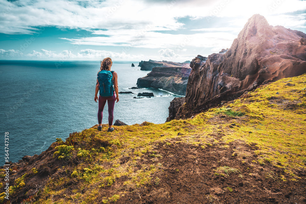 Obraz premium Backpacker woman enjoys panoramic view from a steep cliff overlooking the sea and the rugged foothills of Madeira's coast in the morning. Ponta do Bode, Madeira Island, Portugal, Europe.