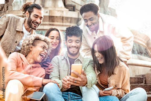 Multiethnic group of smiling people using mobile phone outdoors -  Cheerful guys and girls friends having fun watching at digital display sitting in the city street - Social media lifestyle.