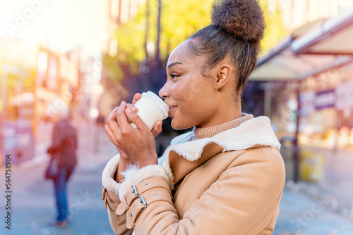 Cheerful African woman holds cup of coffee. A smiling curly brunette lady in brown sweater waiting a tram.