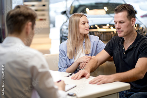 Young couple talking with car dealer in auto salon, man and woman listening to manager while sitting behind table in modern cars showroom. rear view on affable nice car dealer