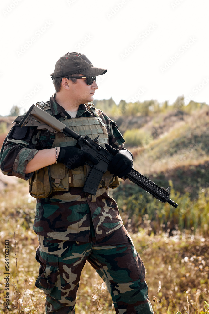 Portrait Of Young Caucasian Man In Military Combat Uniform, Looking At Side Holding Weapon Rifle Gun In Hands, Standing Alone In Field, Ready To Shoot. Military Forces concept