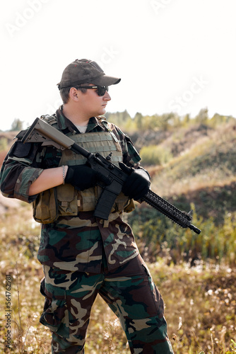 Portrait Of Young Caucasian Man In Military Combat Uniform, Looking At Side Holding Weapon Rifle Gun In Hands, Standing Alone In Field, Ready To Shoot. Military Forces concept