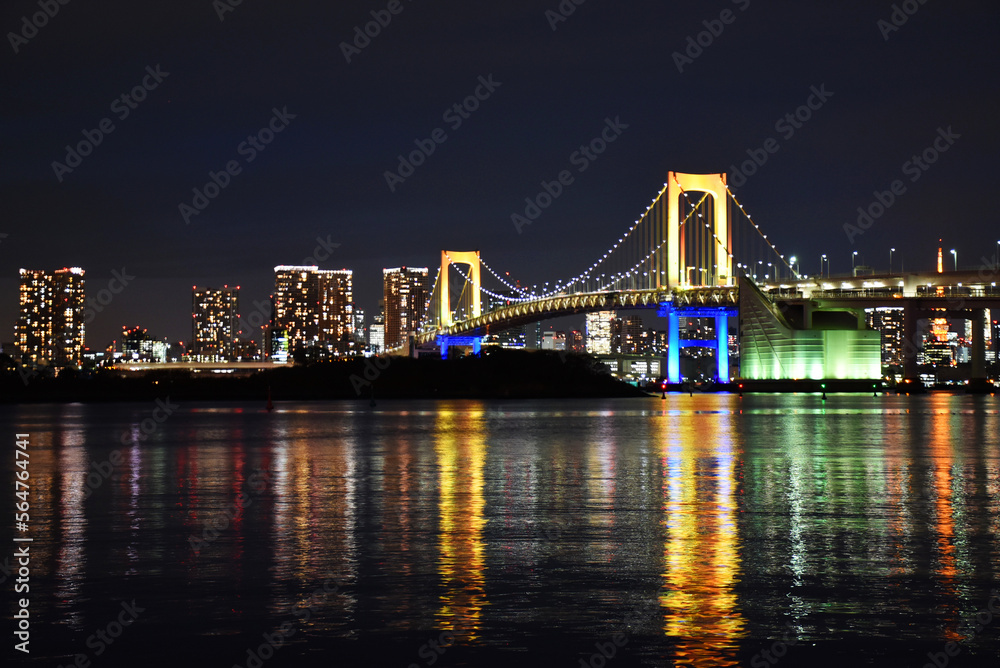 The Rainbow Bridge is a suspension bridge in Odaiba, Tokyo. Illuminated city view with colorful reflection in the water, night view of the Japanese capital city with tall skyscrapers.