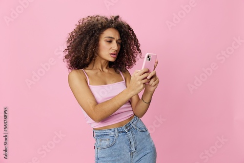 Woman blogger holding phone with curly hair in pink top and jeans poses on pink background, copy space, technology and social media