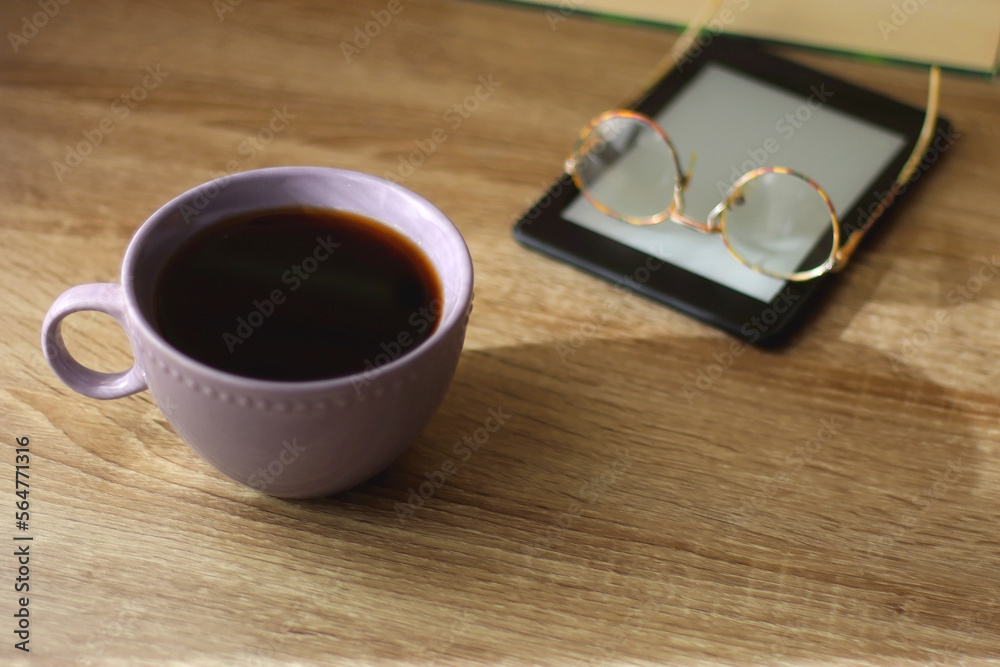 Cup of tea or coffee, e-reader and reading glasses on the table. Pile of books in the background. Selective focus. Cup of tea or coffee, e-reader and reading glasses on the table. Pile of books in the background. Selective focus.