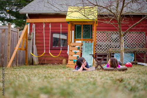 Two children lay in  grass in yard talking with playset in background