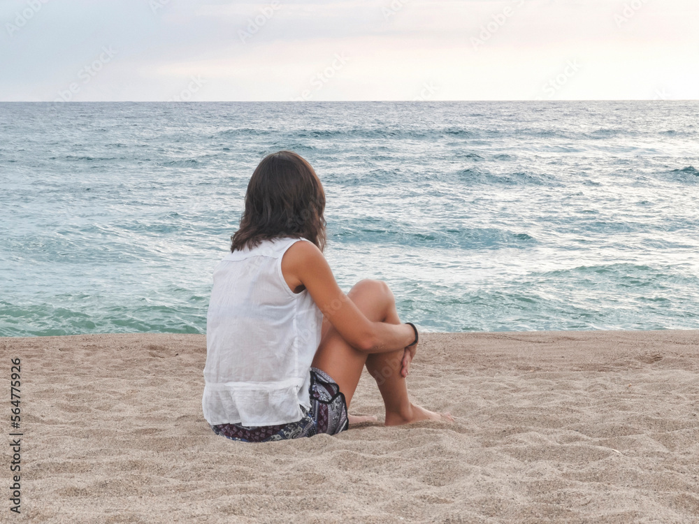 Woman looking at sea while relaxing on sand against sky during sunset Woman looking at sea while relaxing on sand against sky during sunset