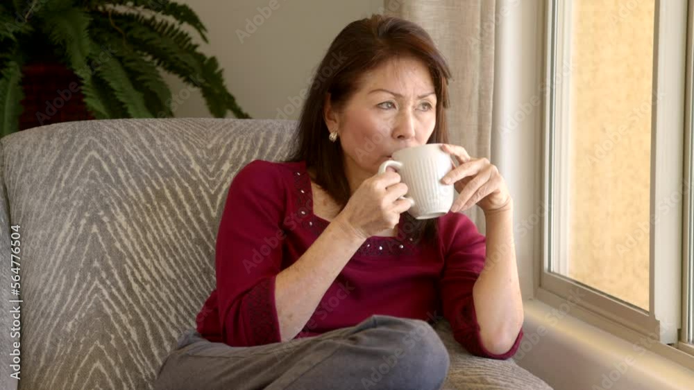 Attractive mature woman drinking coffee relaxing looking out window seated in comfortable chair in home living room Attractive mature woman drinking coffee relaxing looking out window seated in comfortable chair in home living room