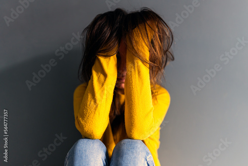 Upset young depress woman in jeans and sweater touching long brown hair while sitting on floor leaning on gray wall