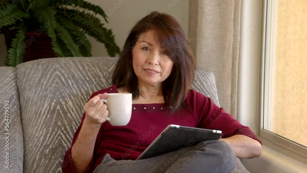 Smiling attractive mature woman looking at camera reading story on mobile tablet drinking coffee relaxing near window in home living room Smiling attractive mature woman looking at camera reading story on mobile tablet drinking coffee relaxing near window in home living room