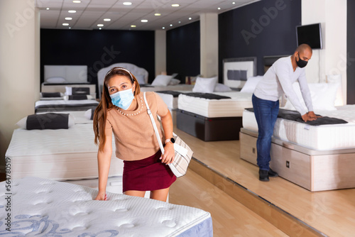 Young cheerful positive Hispanic woman in medical face mask looking for new mattress for bed in furniture store. New lifestyle during coronavirus pandemic