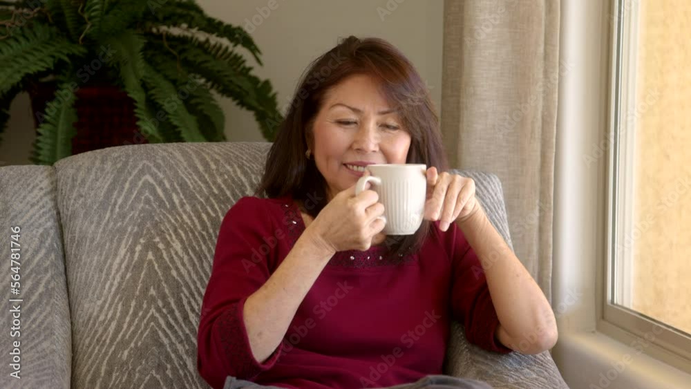 Smiling attractive middle age woman waving looking at camera drinking coffee relaxing near window in home living room Smiling attractive middle age woman waving looking at camera drinking coffee relaxing near window in home living room