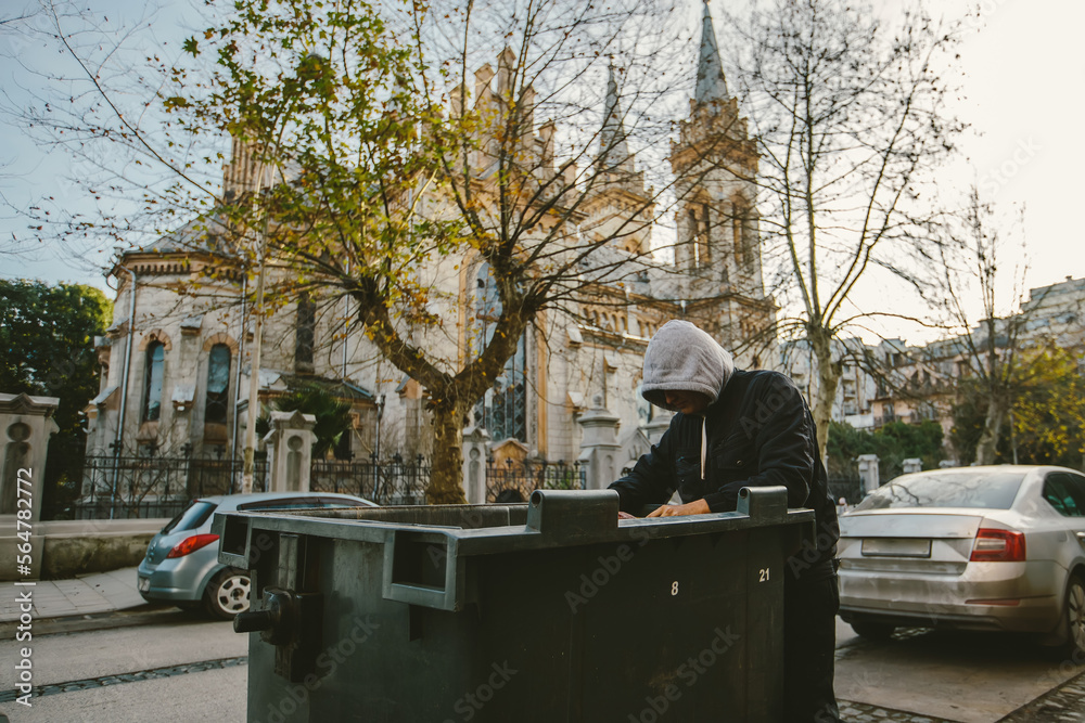 Homeless man looking into trash box in search for food on church background. Homelessness problem concept