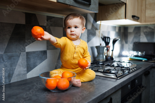 A little boy 1 year old in yellow clothes is sitting in the kitchen on the table with a plate of orange tangerines. Portrait of a cute one-year-old boy and sweet citrus fruits.